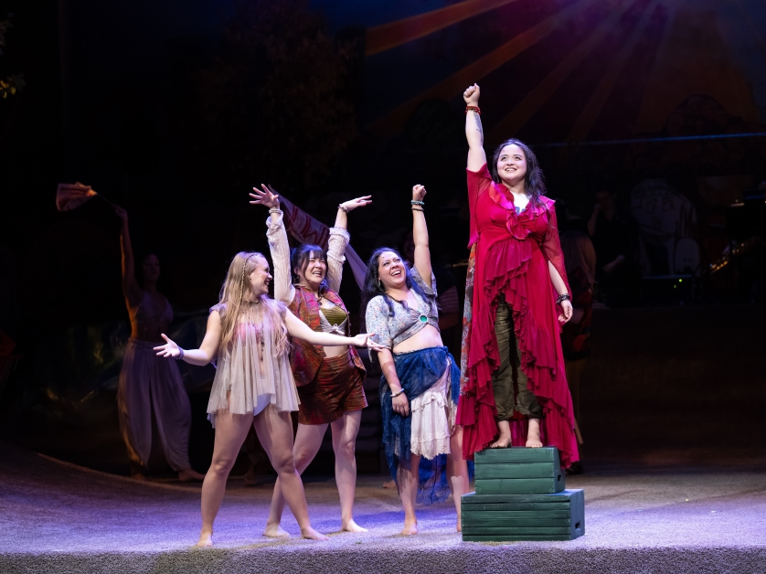 Maddie Tran, a UP alum, stands on a plinth block with her arm raised, with three women standing beside beside her in admiration during a scene from a theatrical production of the musical Hair.
