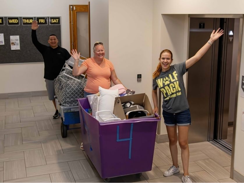 A student and two parents with smiling and waving as they move belongings into the dorm 