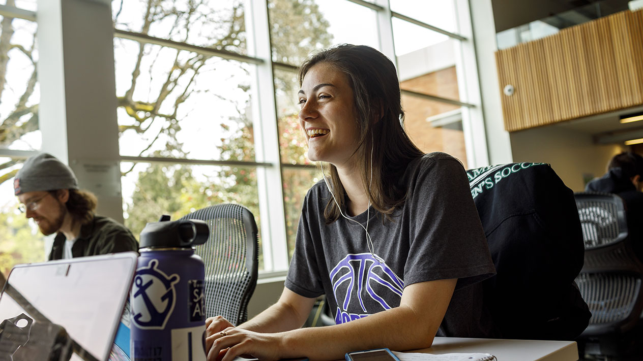 A student smiling while working on their laptop in a common area