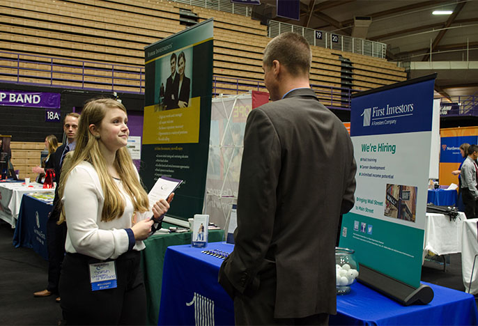 job fair: student holding a resume speaking with an employer in front of a row of tables with posters and information about jobs