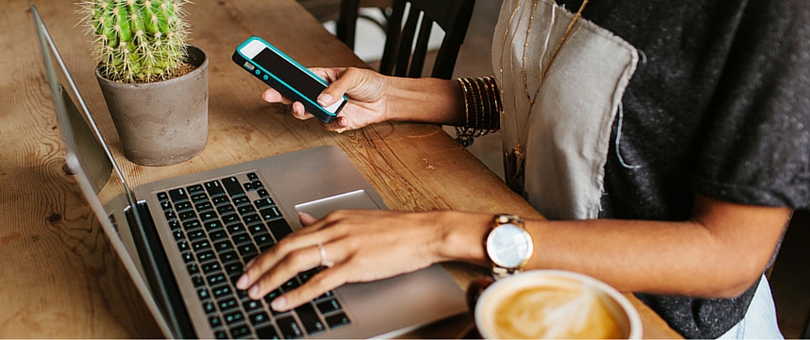 A person typing on a computer while also holding a phone