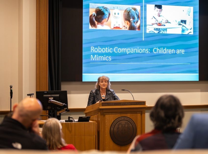 A lecturer in an auditorium speaks in front of a screen displaying a digital presentation