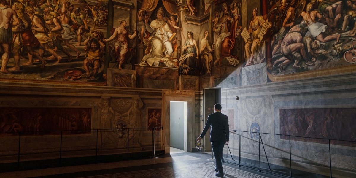 A man in a suit walks away from the camera toward a light filled doorway that pierces an elaborately frescoed wall.