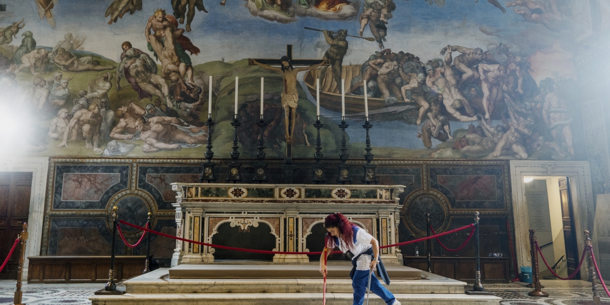 A woman with a dustpan and broom sweeps the floor in front of a large altar in an elaborately frescoed chapel of the Vatican.