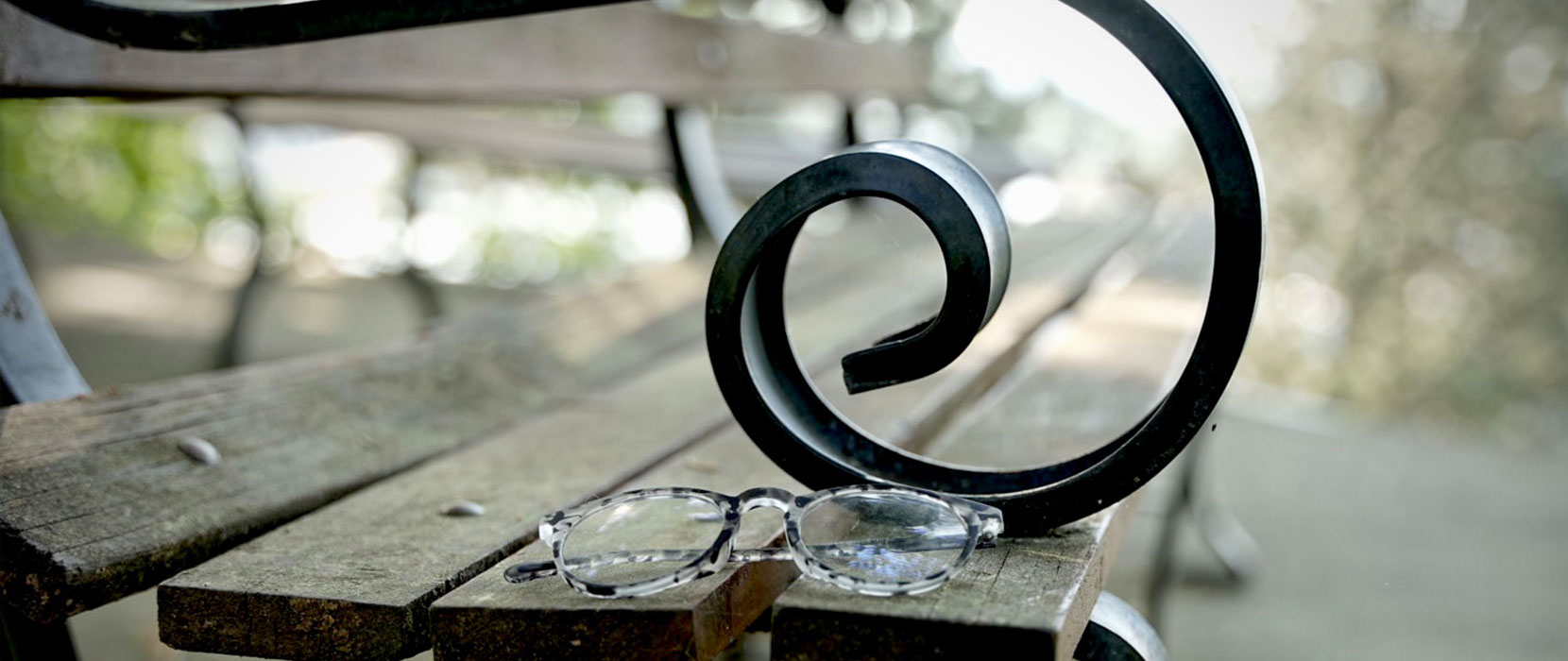 A close-up image of the curved arm of a park bench, upon which sits a pair of glasses, with blurred greenery in the background.