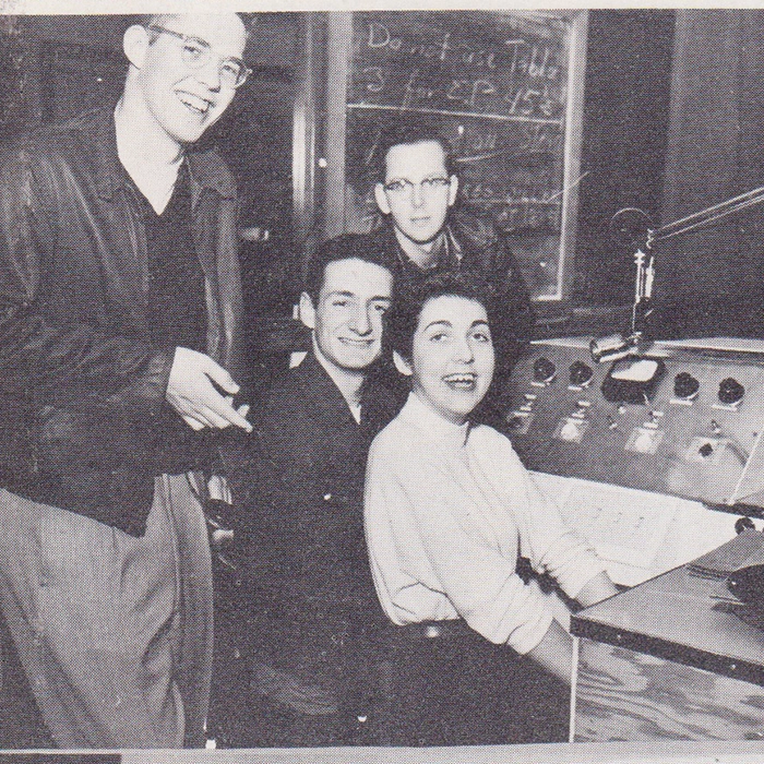 Four people sit at the soundboard of a radio station in a black and white photograph from the 1950s