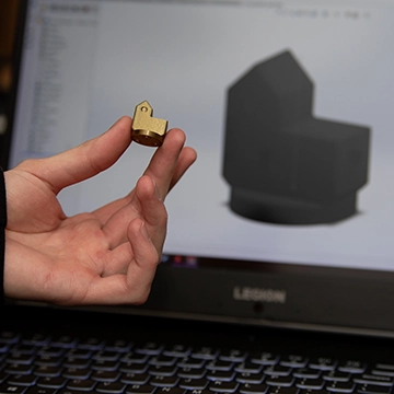 A hand holds a small brass part in front of a computer screen displaying the 3D design of the part