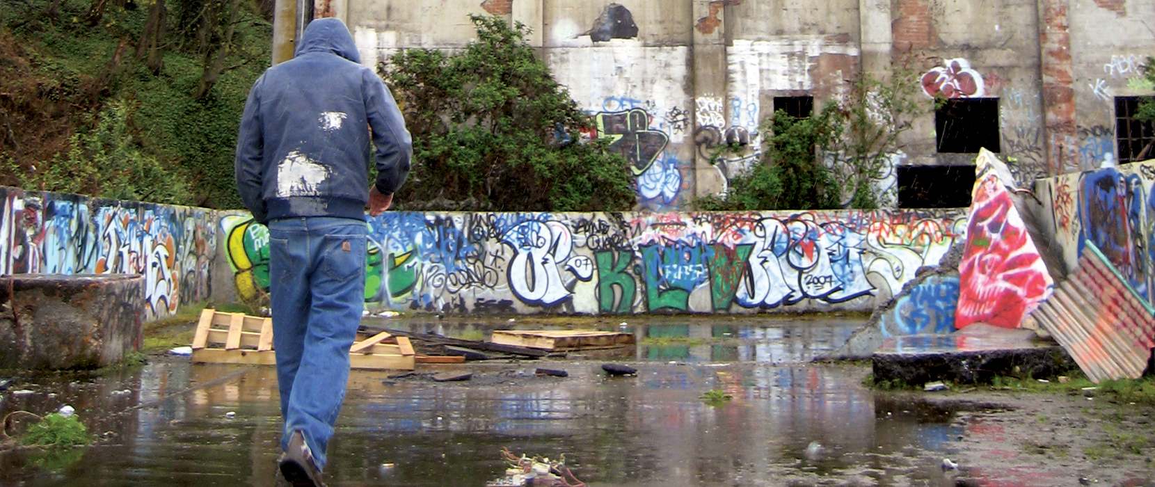 a man walks toward a crumbling industrial building covered in graffiti